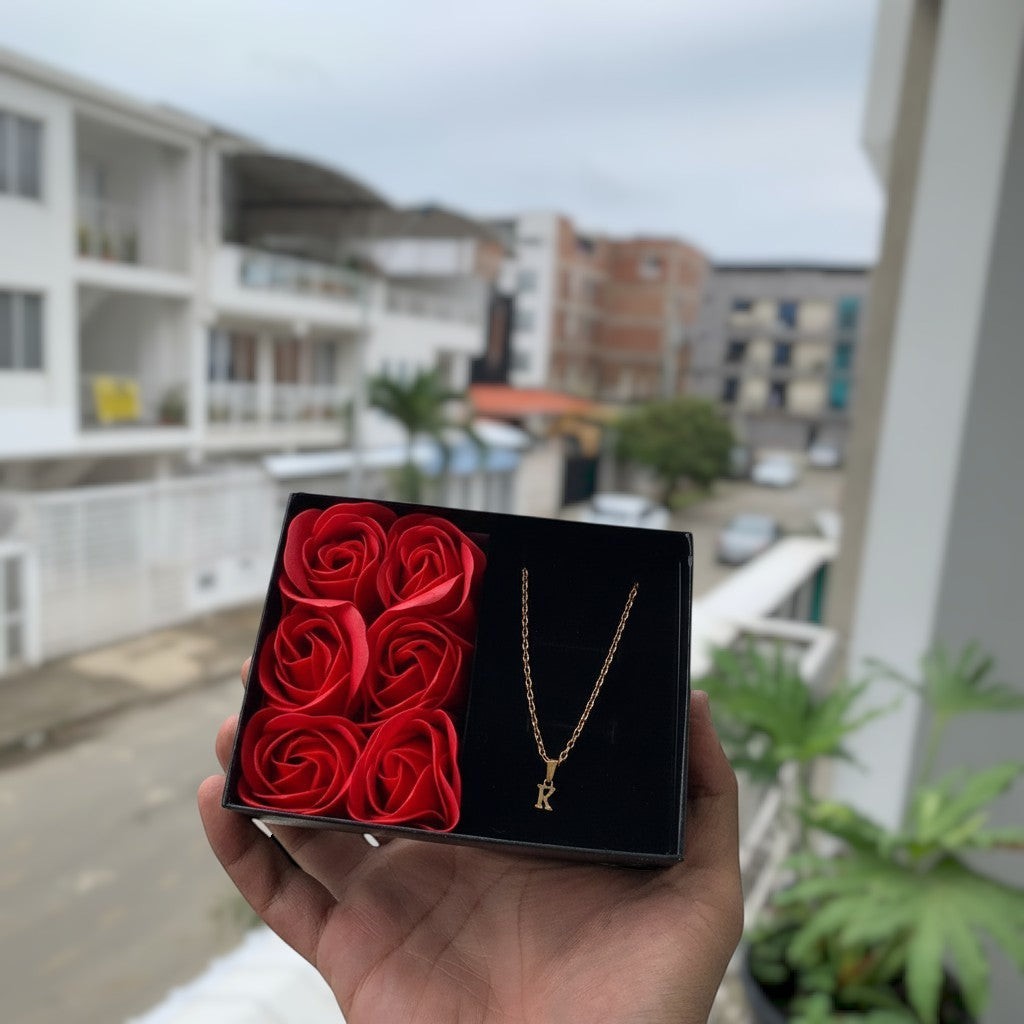 Magillas cadena dorada con colgante de letra K, presentada en caja negra con rosas rojas.