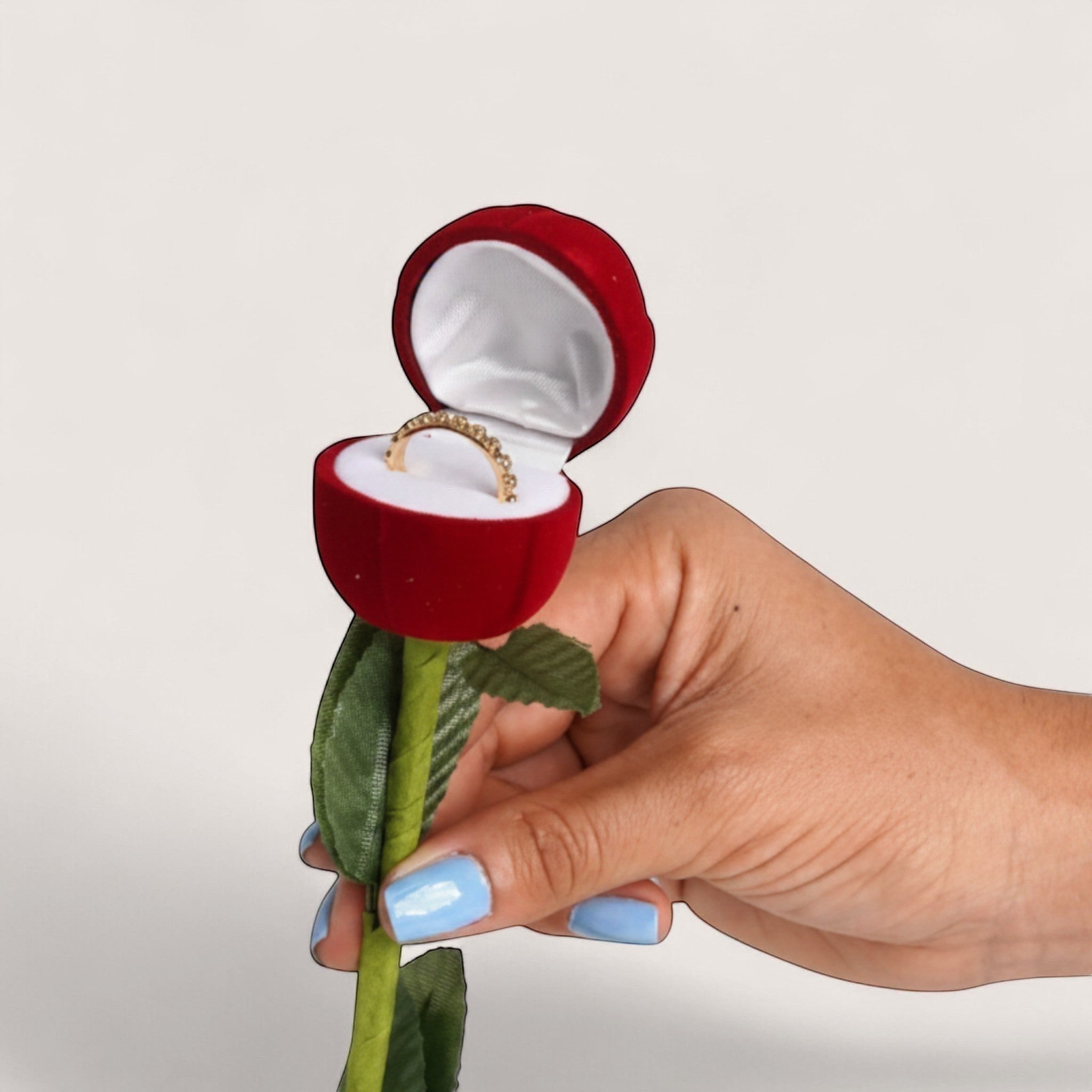 Estuche de terciopelo rojo en forma de rosa con anillo de oro, sostenido por una mano con uñas pintadas de azul claro.
