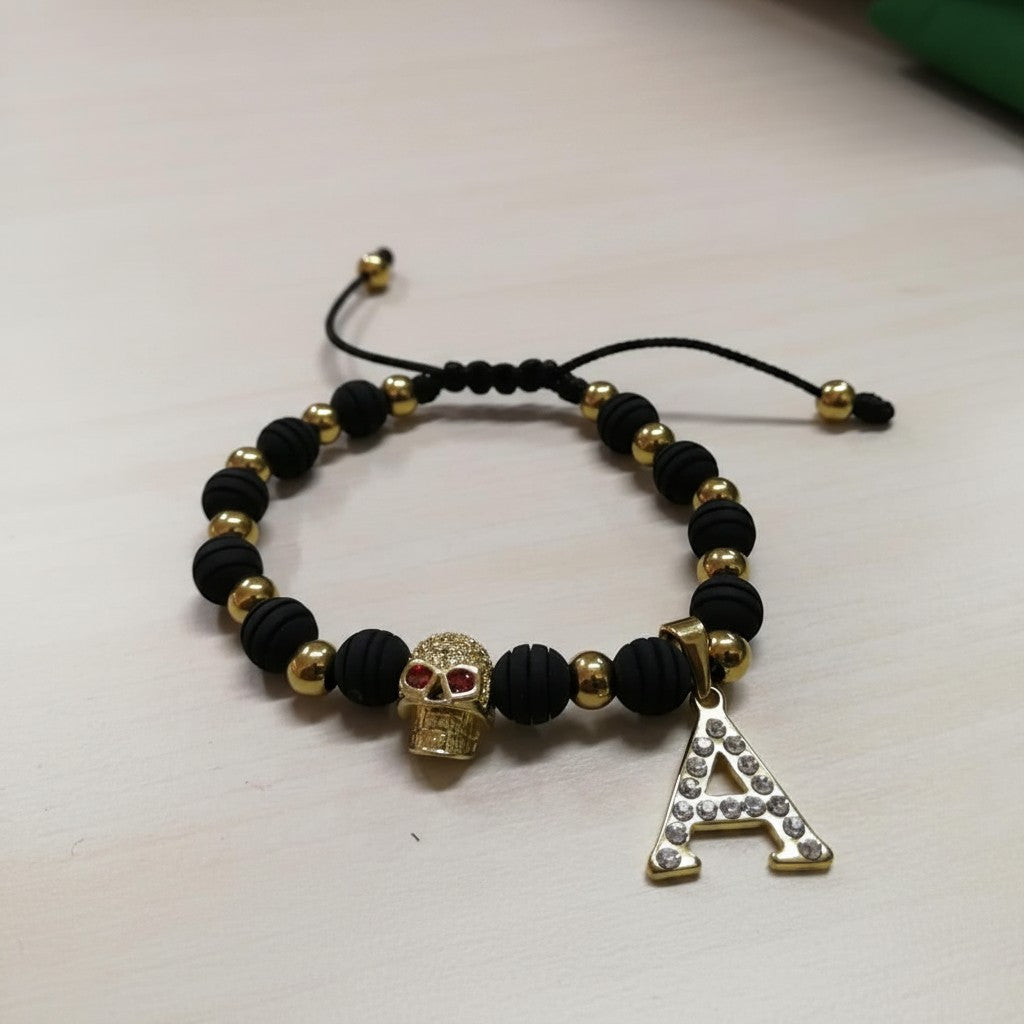Pulsera Magillas de acero inoxidable, neopreno y rodio con cuentas negras y doradas, dije de calavera y letra A con circonitas.