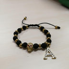 Pulsera Magillas de acero inoxidable, neopreno y rodio con cuentas negras y doradas, dije de calavera y letra A con circonitas.