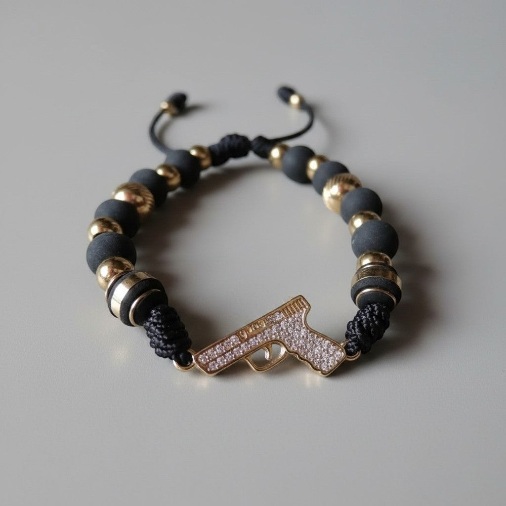 Pulsera Magillas de acero inoxidable, neopreno y rodio con charm de pistola incrustado en circonitas y cuentas negras y doradas.