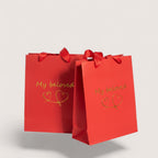 Magillas red paper gift bags with gold My Beloved text and heart illustrations, featuring red ribbon handles.